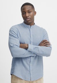 Cămașă albastru deschis, cu mâneci lungi și guler mandarin, realizată dintr-un material texturat, având nasturi albi și o ușoară tăietură la spate.