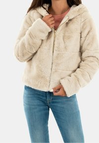Chaqueta de pelaje sintético color crema con capucha, con cierre de cremallera y textura suave. Combinada con una camiseta rosa claro y jeans azules.