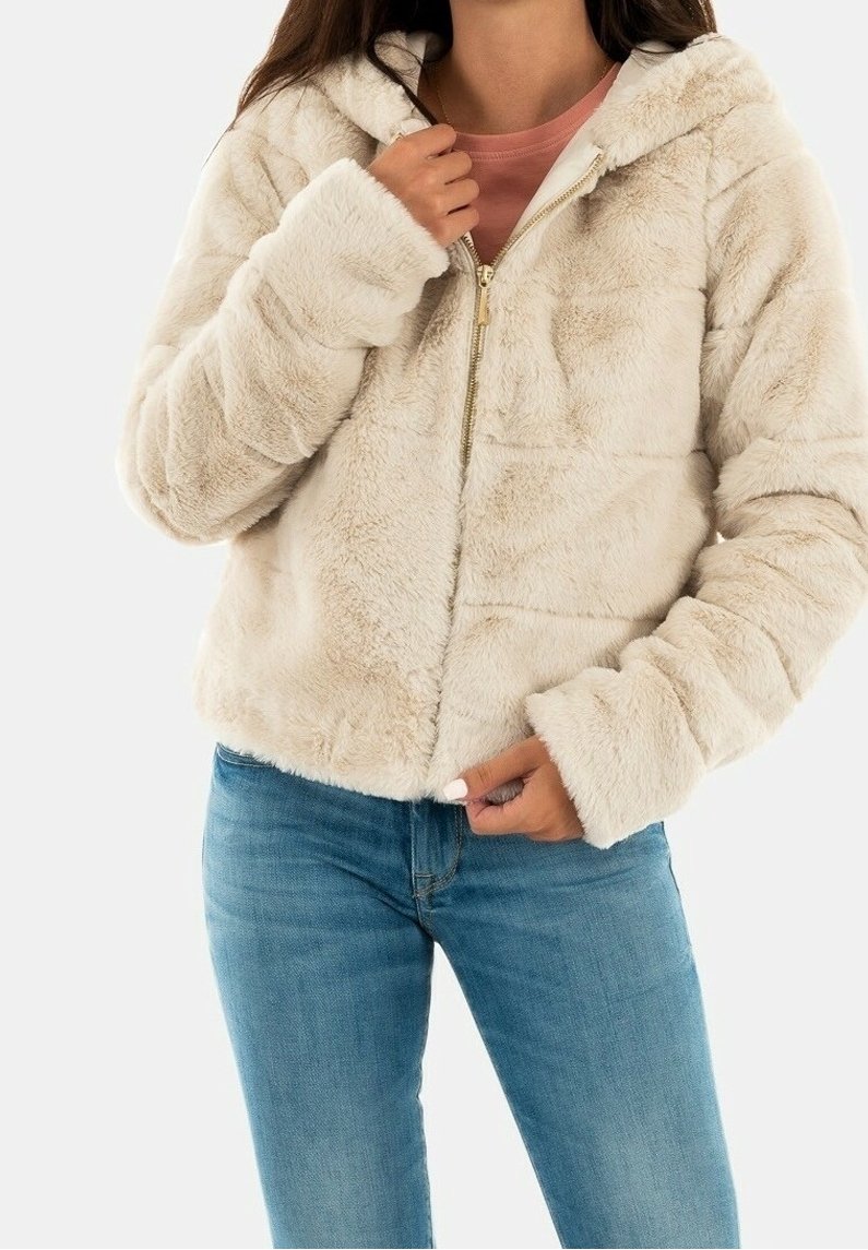 Chaqueta de pelaje sintético color crema con capucha, con cierre de cremallera y textura suave. Combinada con una camiseta rosa claro y jeans azules.