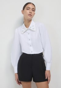Weiße Bluse mit einem großen, verzierten Kragen, der Perlen- und Blumenmuster aufweist, kombiniert mit hochgeschnittenen schwarzen Shorts. Glatte Textur, mit Knopfverschluss vorne.
