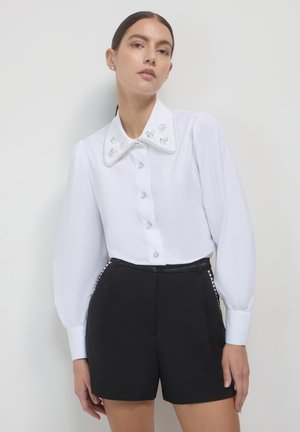 White blouse with a large, embellished collar featuring pearl and flower designs, paired with high-waisted black shorts. Smooth texture, button front.