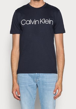 Homme portant un t-shirt noir Calvin Klein et un jean bleu clair, debout devant un fond clair uni.