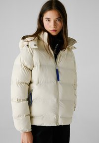 Cremefarbene Pufferjacke mit Reißverschluss vorne, Kapuze und blauen Reißverschluss-Akzenten. Verfügt über ein gestepptes Design und Seitentaschen. Weiche Textur.