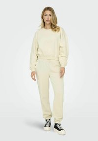 Helles beiges Baumwoll-Sweatshirt und passende Jogginghosen mit Rippbündchen, die einen lockeren Schnitt und minimalistisches Design aufweisen. Schwarze Sneaker mit weißen Sohlen.