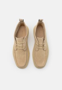 Paire de chaussures en daim beige à lacets avec semelles en crêpe, bouts carrés et languettes de traction marquées à l'arrière sur fond blanc.