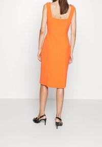 Sukňové oranžové šaty s hranatým výstřihem, silnými ramínky a délkou po kolena. Texturovaná látka se dvěma bočními rozparky. Celkový vzhled doplňují černé sandály na vysokém podpatku.