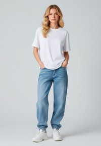 Witte T-shirt met korte mouwen, gecombineerd met lichtblauwe, comfortabele jeans met elastische manchetten en witte sneakers. Neutrale achtergrond.