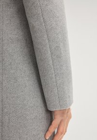 Hand rustend aan de zijkant, gekleed in een lichtgrijze, gestructureerde wollen jas met een diagonaal visgraatpatroon.