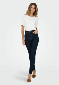 Witte kortaf shirt gecombineerd met high-waisted donkere denim jeans. De jeans hebben lichte stiksels, en de outfit wordt aangevuld met beige hakken.