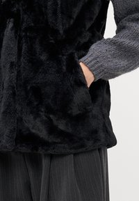 Gilet in ecopelliccia nera con una texture morbida e una vestibilità rilassata, abbinato a un maglione grigio lavorato a maglia, con due tasche laterali in evidenza.