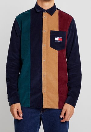 Mand iført en langærmet kordfløjls skjorte med lodrette striber i grøn, navy, tan og bordeaux, med en navy lomme med Tommy Jeans-logo.