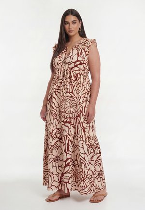 Kvinde med langt mørkt hår bærer en ærmeløs beige maxikjole med brunt blomsterprint og brune flade sandaler, står med den ene hånd ned langs siden.