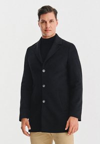 Czarny wełniany płaszcz z wyłogiem z nacięciem, trzema dużymi guzikami i bocznymi kieszeniami. Tkanina jest gładka, a krój dopasowany.