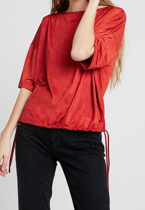 Femme portant une blouse rouge ample avec un cordon à l'ourlet et un pantalon noir, posant avec un bras levé près du visage.