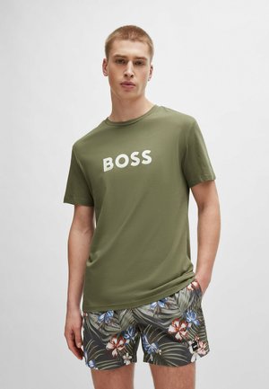 T-shirt in cotone verde oliva con testo bianco "BOSS", abbinato a pantaloni corti neri con un motivo floreale colorato e accenti di fogliame verde.