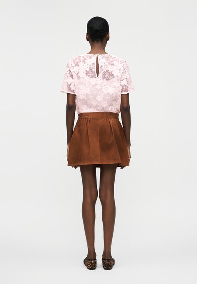 Γυναίκα με κοντά μαύρα μαλλιά που φοράει διάφανο ροζ τοπ από δαντέλα με λουλούδια, καφέ φούστα από σουέτ και φλατ παπούτσια με λεοπάρ σχέδιο, στέκεται γυρισμένη με τη πλάτη.