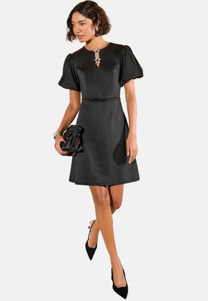 Schwarzes Kleid mit kurzen Puffärmeln, subtiler Taillennaht und sternförmigen Verzierungen. Begleitet von einer schwarzen Clutch und spitzen Absätzen.
