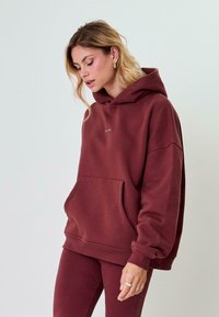 Bordó, oversized kapucnis pulóver, puha anyagból készült, elöl zsebbel, bordázott mandzsettával és egy egyszerű logóval a mellrészen.