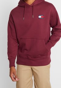 Bordowa bluza z kapturem, z trokami i kieszonką z przodu. Posiada małe białe i niebieskie logo Tommy Jeans na piersi. Miękka faktura, luźny krój.