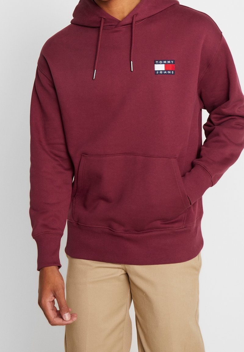 Bordowa bluza z kapturem, z trokami i kieszonką z przodu. Posiada małe białe i niebieskie logo Tommy Jeans na piersi. Miękka faktura, luźny krój.