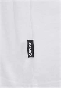 Hvit bomulls T-skjorte med et vevd svart merke som har teksten "CAYLER." plassert nær kanten. Glatt tekstur og enkelt design.