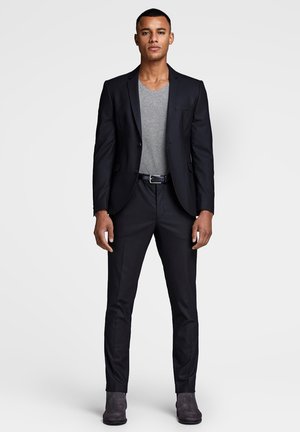 Black suit jacket over a grey v-neck shirt, paired with tailored black trousers and grey suede boots, showcasing a fitted design.
