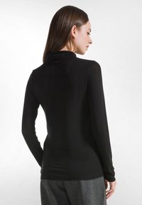 Schwarzes Langarm-Turtleneck-Oberteil aus glattem, figurbetontem Stoff, mit einem subtilen Glanz und hohem Kragen. Kombiniert mit grau gestreiften Hosen.