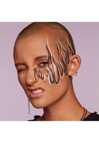 Personne chauve avec un maquillage facial comportant des designs de flammes en brun, jaune et blanc sur la joue droite, portant des boucles d'oreilles en forme de créoles et un haut noir.