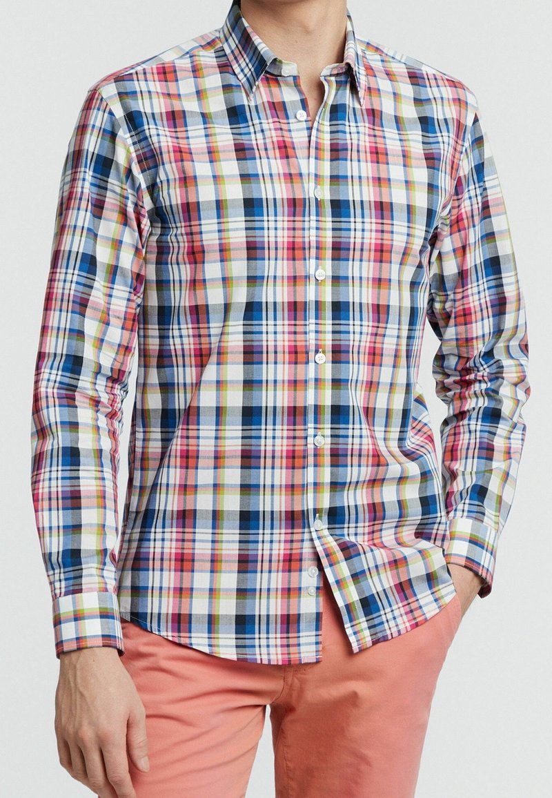 Homme portant une chemise à manches longues boutonnée à carreaux multicolores et un pantalon corail, une main dans la poche, sur un fond uni.