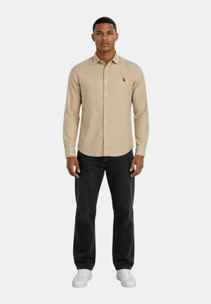 Mann steht frontal, trägt beige langärmliges Hemd mit Knöpfen, schwarze Jeans und weiße Turnschuhe vor einem schlichten weißen Hintergrund.