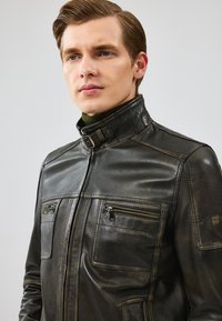 Veste en cuir noir avec un col haut, un devant zippé et plusieurs poches. Présente une finition texturée et des accents de couture contrastante.