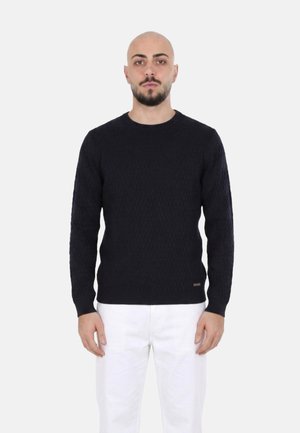 Uomo con testa rasata e barba, che indossa un maglione nero lavorato a treccia e pantaloni bianchi, in piedi davanti a uno sfondo chiaro uniforme.