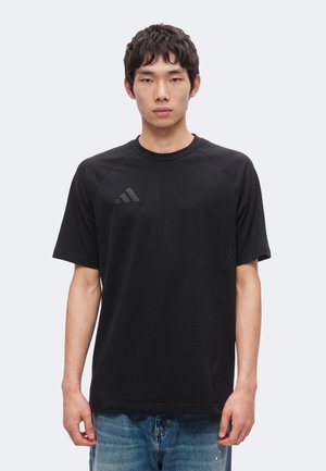 Ung mand iført en ensfarvet sort t-shirt med korte ærmer og et diskret logo på brystet samt blå jeans, set forfra med et neutralt ansigtsudtryk.