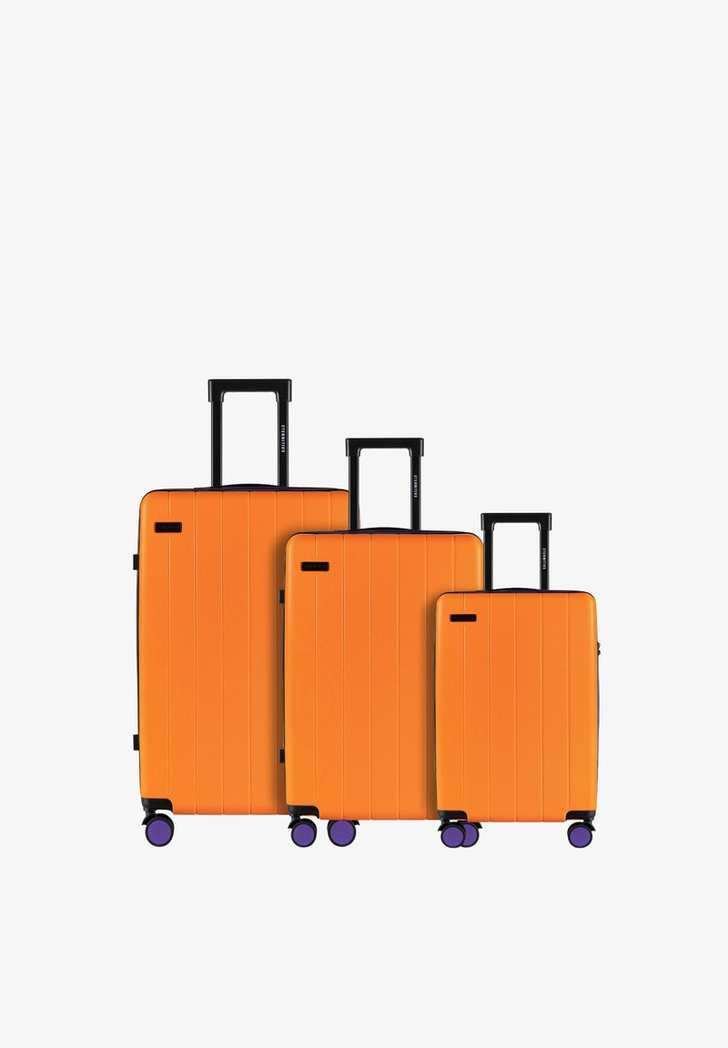 Trzy pomarańczowe walizki z twardą powłoką o różnych rozmiarach, z pionowymi rowkami, czarnymi wysuwanymi rączkami i fioletowymi kółkami.