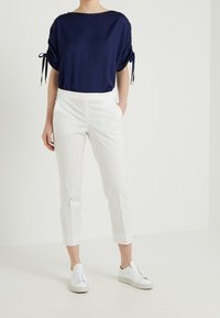 Person wearing a navy blue short-sleeve top with tied sleeves, white cropped pants, and white lace-up sneakers, standing with one hand in pocket.