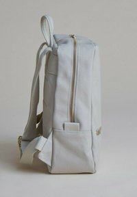 Ljusgrå ryggsäck med ett texturerat tyg, guldfärgad dragkedja, justerbara remmar och sidoficka. Kompakt fyrkantig form, minimalistisk design.