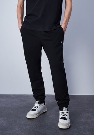 Pantaloni jogger neri in cotone con vita elastica, vestibilità slim e un piccolo logo di lato, abbinati a sneaker bianche e grigie.