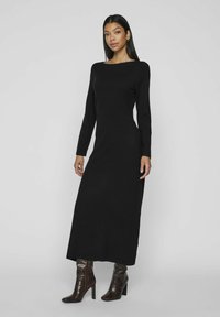 Abito maxi nero a maniche lunghe con scollatura a barca, realizzato in tessuto morbido, caratterizzato da una silhouette aderente e uno spacco laterale. Indossato con stivaletti alla caviglia con motivo.
