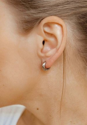 Silberne Creolen mit einer glatten, glänzenden Oberfläche und einem leicht gedrehten Design. Getragen im durchstochenen Ohr, nah an heller Haut.