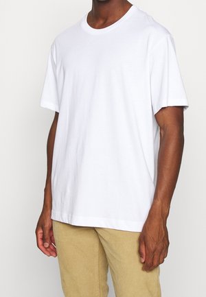 Uomo con maglietta bianca a maniche corte semplice e pantaloni beige chiaro, in piedi davanti a uno sfondo bianco, braccia rilassate lungo i fianchi.