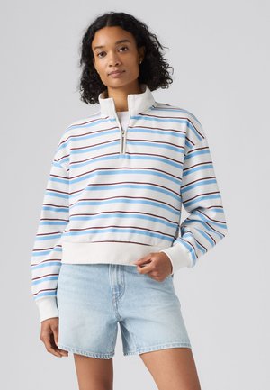 Femme portant un pull blanc avec des rayures horizontales bleues et marron et un short en jean bleu clair, debout devant un fond uni.