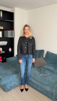Kvinde med blondt hår iført en sort quiltet jakke, blå jeans og sorte spidse sko, der står ved siden af en grønblå veloursofa med puder.