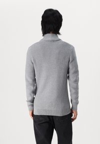 Grå stickad tröja med hög krage, texturerat mönster och ribbad fåll, i kombination med svarta byxor. Enbart bakvy.