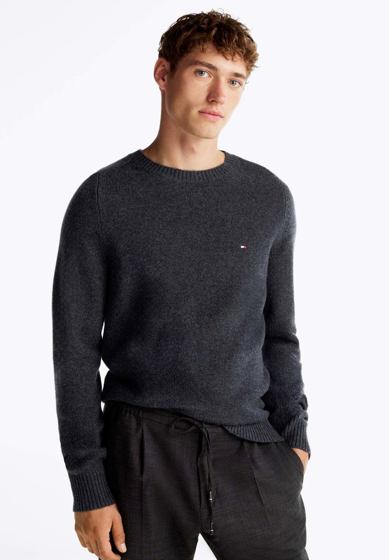 Maglione lavorato a maglia grigio scuro con scollo rotondo e polsini a costine, caratterizzato da un piccolo logo sul petto. Il materiale sembra morbido e strutturato.