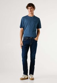 Modrá bavlnená tričko s okrúhlym výstrihom, spárované s tmavými džínsami. Topánky majú krémovú farbu s hnedými akcentmi a svetlými šnúrkami.