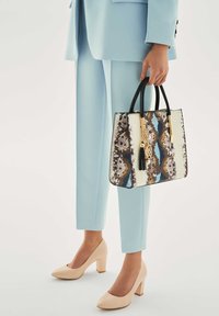 Rankų darbo krepšys su roplio odos raštu, juodomis rankenomis, kremine ir ruda detalėmis, puoštas auksine pakabuka ir kutu. Dėvimas su šviesiai mėlynu kostiumu ir smėlio spalvos aukštakulniais.