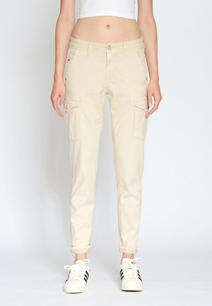 Femme portant un pantalon cargo beige avec des poches à rabat à l'avant et des baskets blanches à rayures noires, debout devant un fond uni.