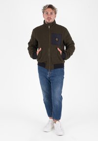 Giacca bomber verde oliva con colletto alto, chiusura a zip, tasca frontale con zip di colore navy e polsini a coste, abbinata a jeans blu e sneakers bianche.