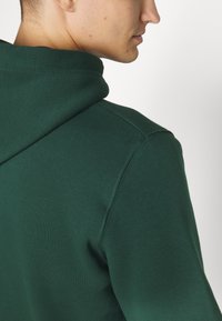Dark green hoodie with a soft, textured fabric. Features a drawstring hood and reinforced shoulder seams, showcasing a relaxed fit.