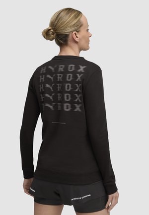 Sudadera negra con la palabra "HYROX" impresa en diferentes opacidades en la parte de atrás. Material suave con puños y dobladillo acanalados, que complementa pantalones cortos atléticos.